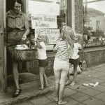 Nainen astuu ulos kaupan ovesta ostoskori kädessään. Kolme kesäasuista lasta pyörii naisen ympärillä. Yksi lapsista osoittaa suoraan kameraan.